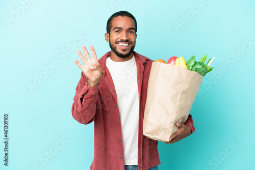 Young latin man holding a grocery shopping bag isolated on blue background happy and counting four with fingers