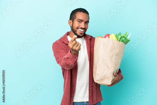 Young latin man holding a grocery shopping bag isolated on blue background making money gesture