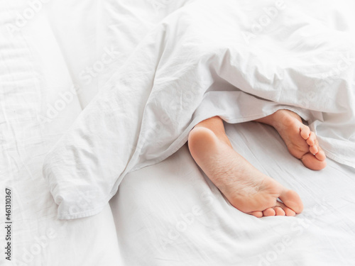 View on bare feet under blanket. Bedroom lit with morning light. Woman sleeps with comfort. White crumpled linen.