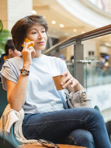 Woman is talking by smartphone and drinking coffee at shopping mall. Coffee break at store.