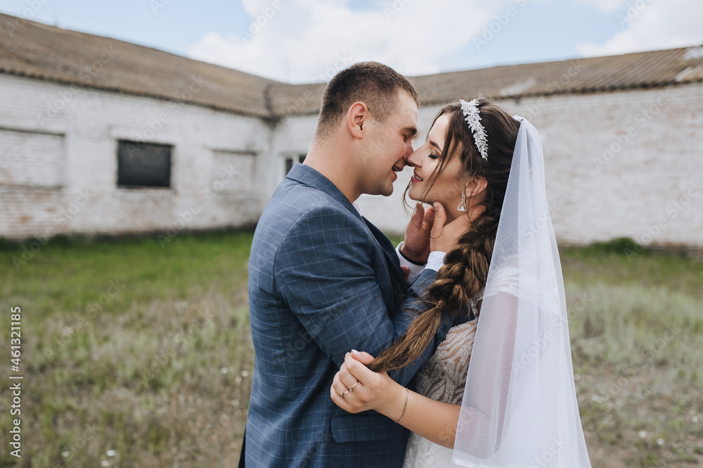 Smiling groom in a blue suit and a brunette bride in a white dress with a pigtail are hugging, standing in a village in nature on the background of a brick building. Close-up wedding photography.