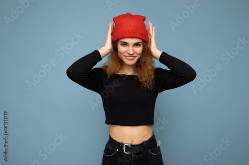 Shot of young nice winsome brunet woman wavy-haired with sincere emotions wearing black crop top and red hat isolated on blue background with free space and covering ears with hands