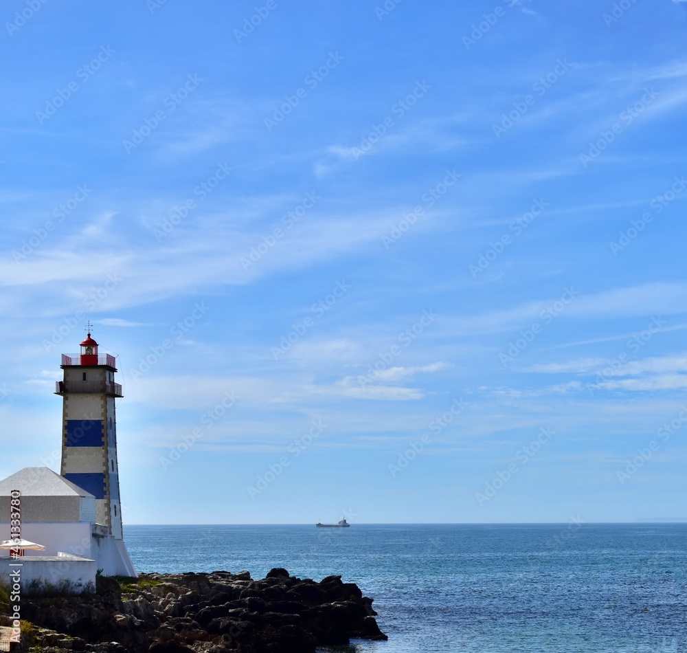 Lighthouse tower on background of blue sky and blue sea with  small ship silhouette on horizon