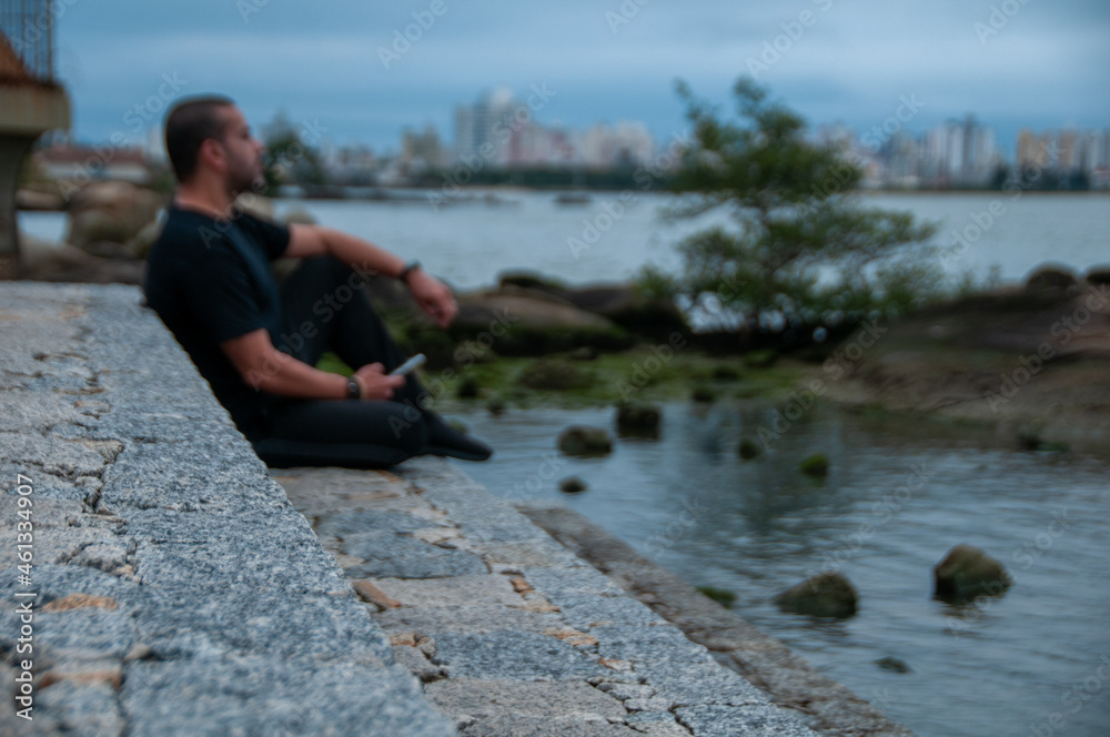 man in blurred image wearing dark clothes sitting on the rock in front of the beach in cloudy afternoon with cell phone in his hands thoughtful, landscape in the background. man in blurred image wearing dark clothes sitting on the rock in front of the beach in cloudy afternoon with cell phone in his hands thoughtful, landscape in the background.