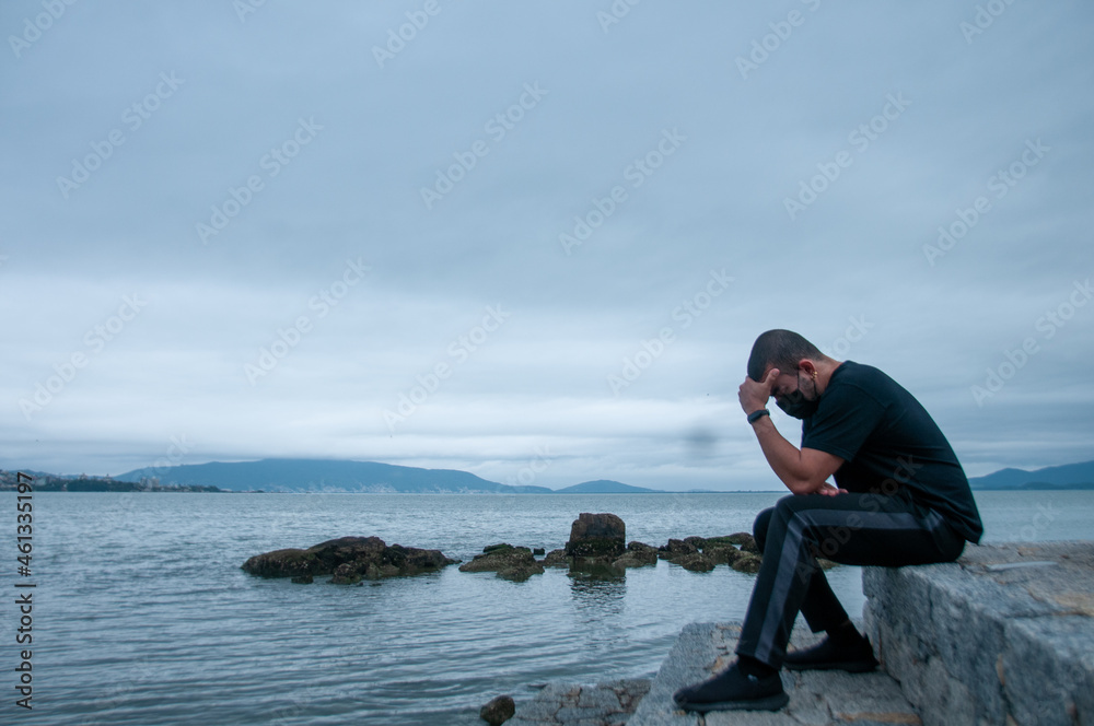 man wearing dark clothes, mask, pensive in a cloudy afternoon, sitting on the beach with his hands on his head, in the background gray sky and landscape of rocks and sea water