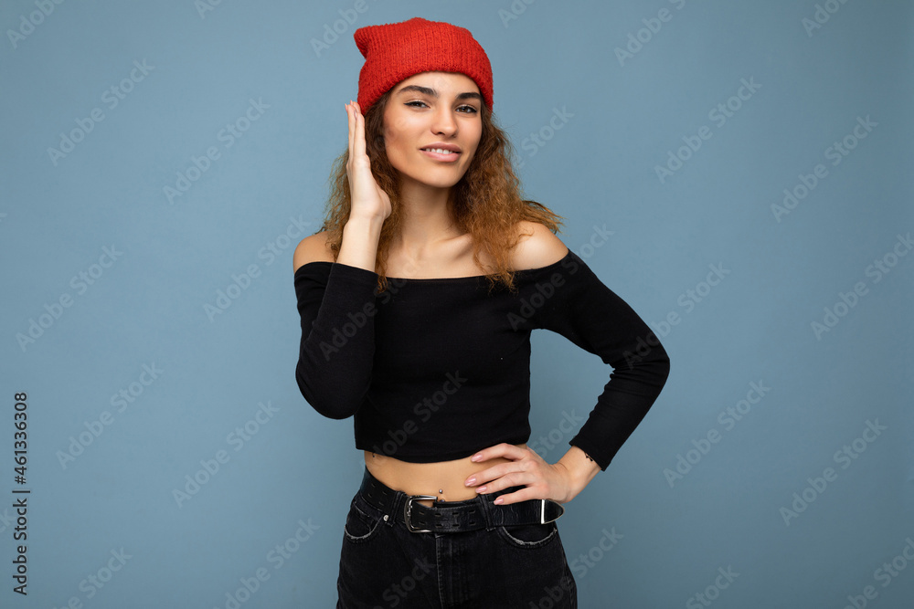 Photo of young positive happy beautiful brunette curly woman with sincere emotions wearing black crop top and red hat isolated on blue background with copy space and listening attenively Photo of young positive happy beautiful brunette curly woman with sincere emotions wearing black crop top and red hat isolated on blue background with copy space and listening attenively
