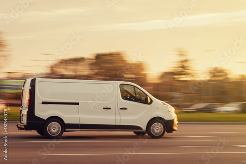 A white van drives down the street towards the sunrise. Commercial transport and small cargo transportation, delivery of goods. Motion blur