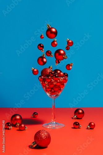 Martini glass with Xmas ornament splashing bubbles against two tone red and blue vibrant background. Christmas or New Year celebration toast. Creative drink concept. Minimal December festive idea.