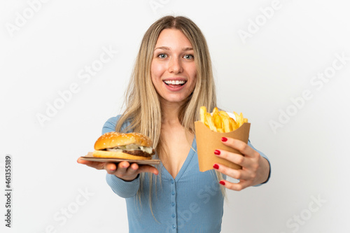 Young blonde woman holding fried chips and cheeseburger over isolated background