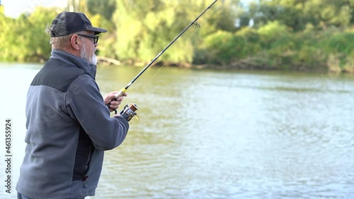 Mature gray-bearded man with glasses fisherman holding reel rod, spinning or spoon-bait and fishing on good summer day. Mens hobby