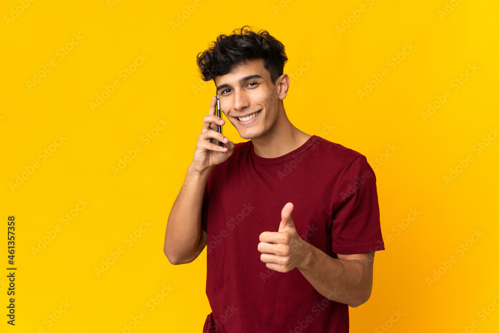 Young Argentinian man isolated on yellow background keeping a conversation with the mobile while doing thumbs up Young Argentinian man isolated on yellow background keeping a conversation with the mobile while doing thumbs up