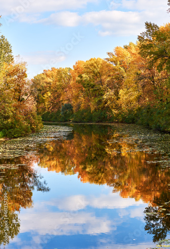 Colorful autumn trees on river banks. Multicolored bright foliage of trees and blue sky reflected in water surface