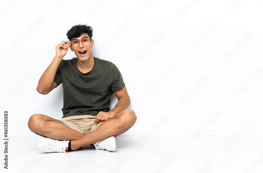 Young Argentinian man sitting on the floor with glasses and surprised Young Argentinian man sitting on the floor with glasses and surprised