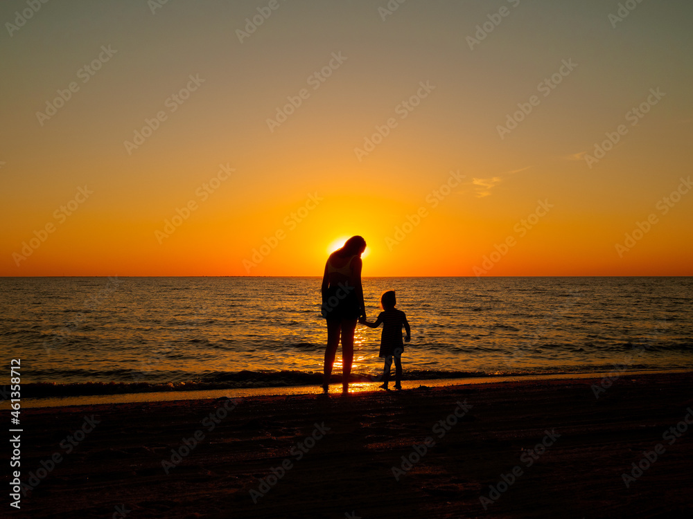 A young woman and a little girl stand on the sea beach and hold hands. In the background, the evening sun setting over the horizon.