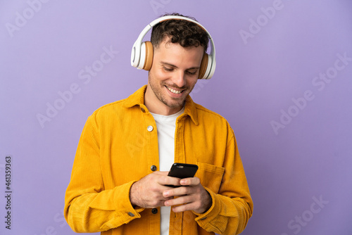 Young caucasian handsome man isolated on purple background listening music and looking to mobile © luismolinero