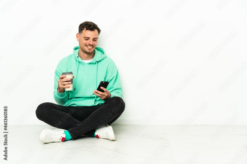 Young handsome caucasian man sitting on the floor holding coffee to take away and a mobile Young handsome caucasian man sitting on the floor holding coffee to take away and a mobile