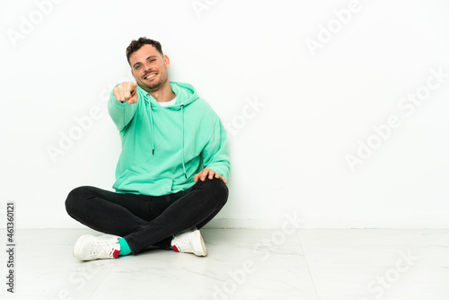 Young handsome caucasian man sitting on the floor points finger at you with a confident expression