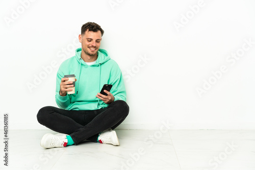 Young handsome caucasian man sitting on the floor holding coffee to take away and a mobile