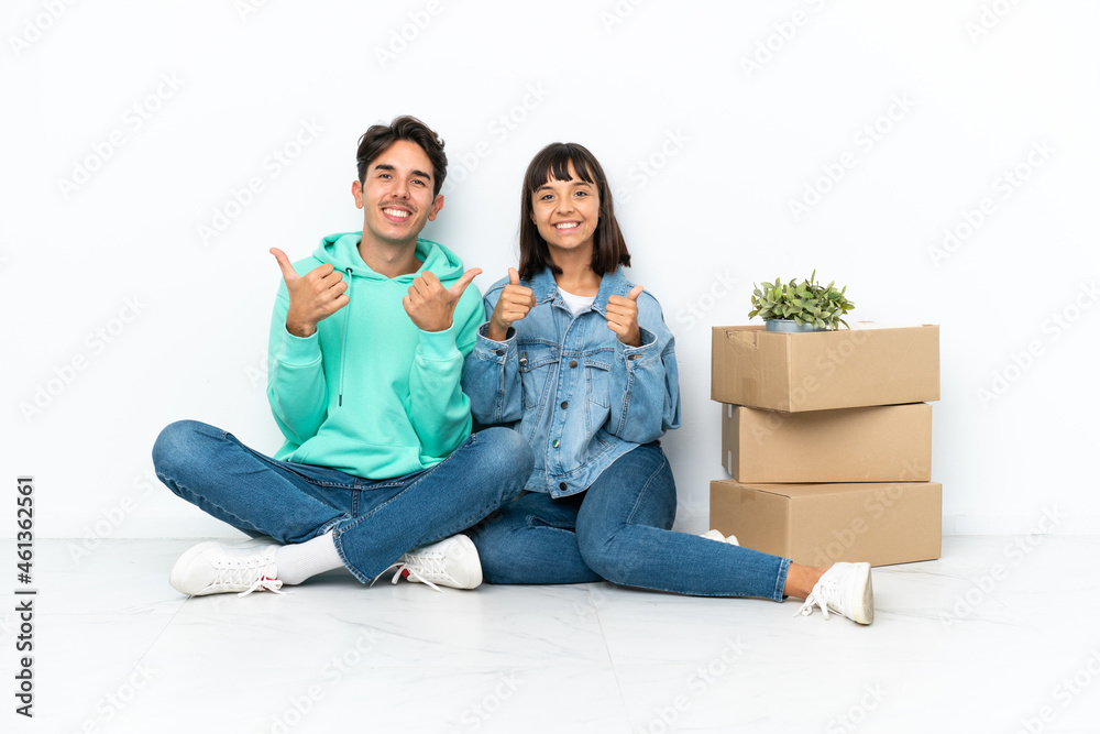 Young couple making a move while picking up a box full of things sitting on the floor isolated on white background giving a thumbs up gesture with both hands and smiling Young couple making a move while picking up a box full of things sitting on the floor isolated on white background giving a thumbs up gesture with both hands and smiling
