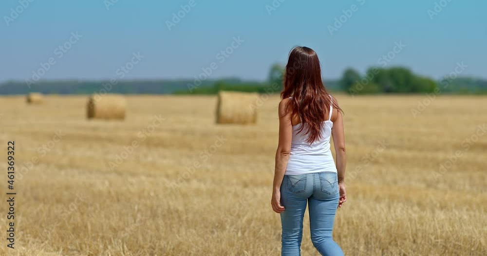 Slender woman in jeans is walking along a mown field, a walk. Large haystacks lie in a golden field, a beautiful rural landscape. Rear view. 4k, ProRes Slender woman in jeans is walking along a mown field, a walk. Large haystacks lie in a golden field, a beautiful rural landscape. Rear view. 4k, ProRes