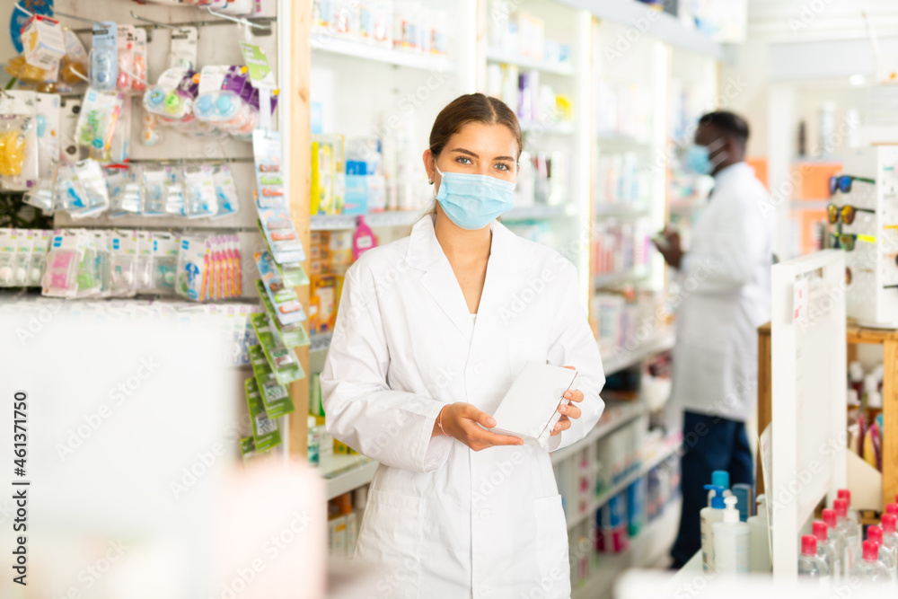 Pharmacist in mask holding drug package in hands. Her colleague working behind.