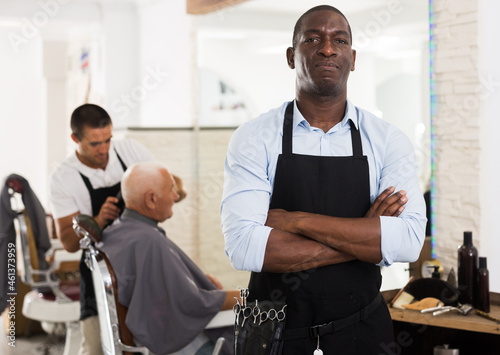Confident African-American man hairdresser posing with folded arms in modern barber shop..