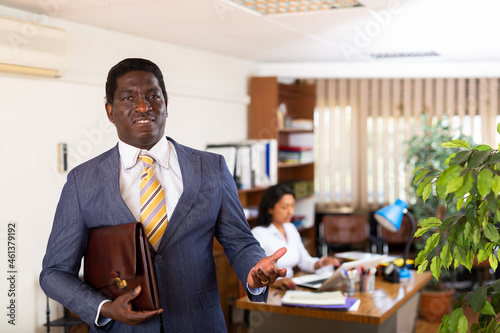 Confident smiling African American man in business suit standing in modern office making inviting gesture with hand. Business partnership invitation concept