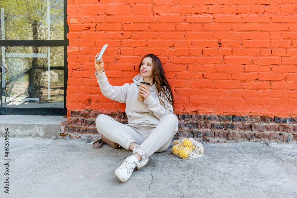 Diversity adult woman, sits on street and drinks coffee from a thermo mug, looks into phone camera and takes selfie.Zero waste.