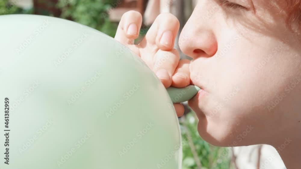 Teenager boy is blowing air balloon. Handsome redhead boy holding balloon in hands inflating green balloon. Mouth closeup. Happy birthday party, celebration.