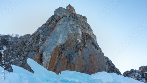 A picturesque granite rock, devoid of vegetation, against a clear azure sky. Steep gray-brown slopes, bizarre outlines. At the base are blocks of blue ice hummocks. Baikal