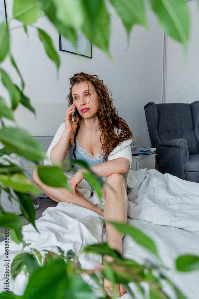 Obraz premium Young woman in comfortable blue pajamas works and talks a cell phone while lying the bed in the bedroom.
