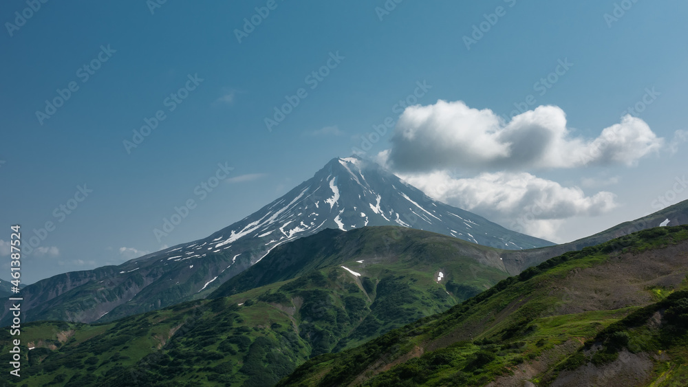 A beautiful conical volcano with snow-covered slopes against a blue sky background. Cumulus clouds near the top. In the foreground are green hills. Kamchatka. Stratovolcano Vilyuchinsky
