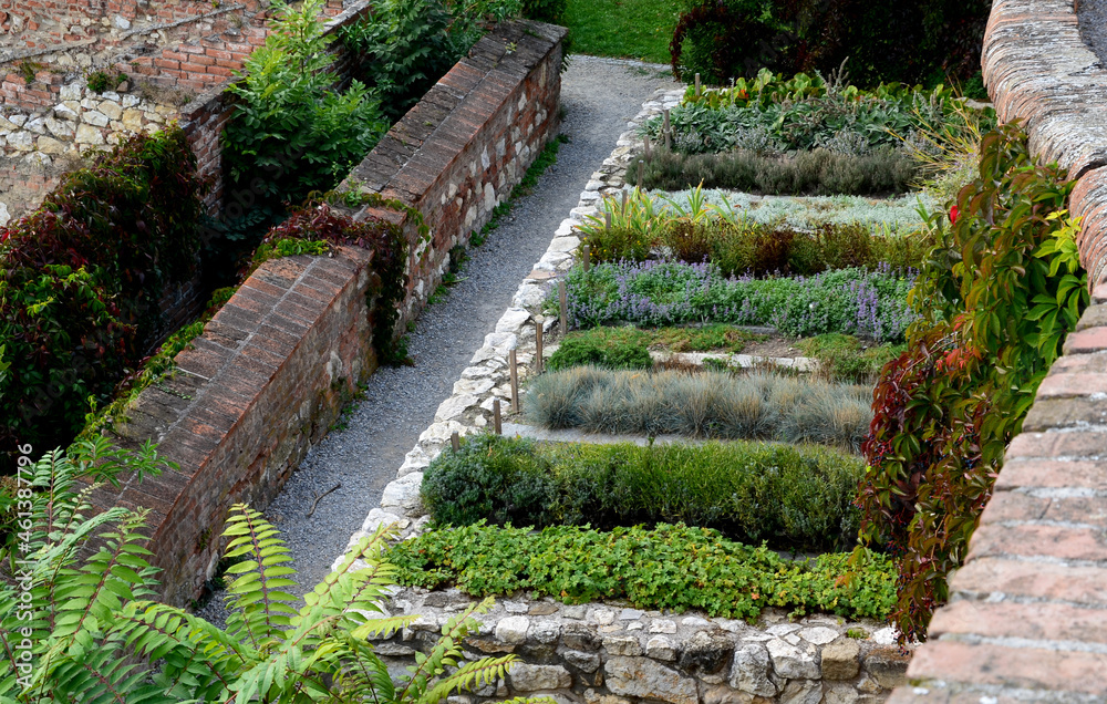 herb beds in rows above a limestone stone back, a brick walled courtyard with a gravel path in the park. monastic healing garden with a focus on the blind. the blind try scents and textures. 