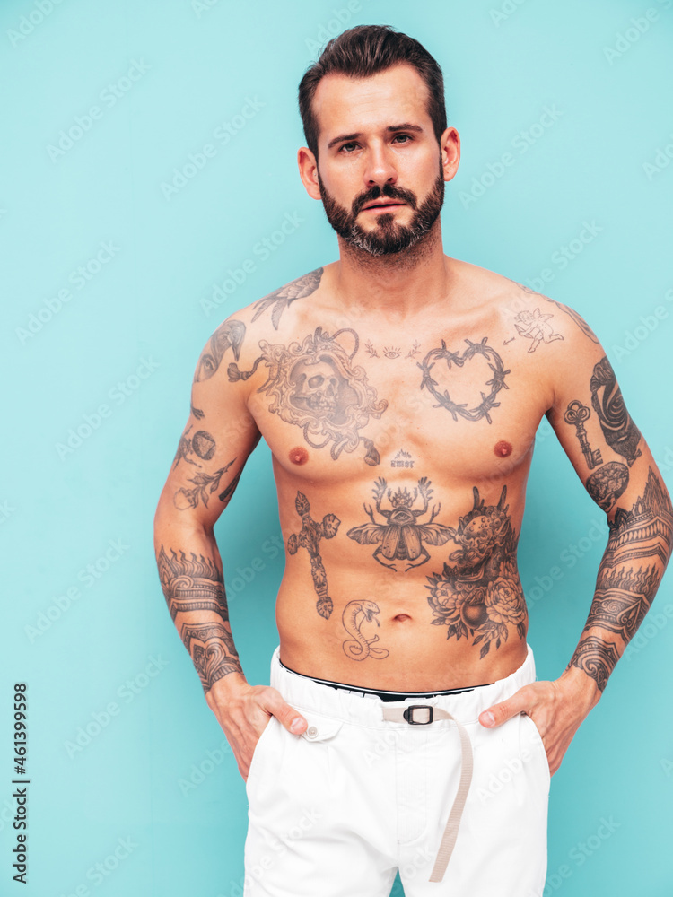 Fototapeta premium Portrait of handsome confident stylish hipster lambersexual model. Sexy modern man. Naked torso with tattoos.Fashion male posing in studio near light blue wall