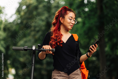 business asian woman , wearing glasses standing with electric scooter outdoors in park, woman using mobile phone photo
