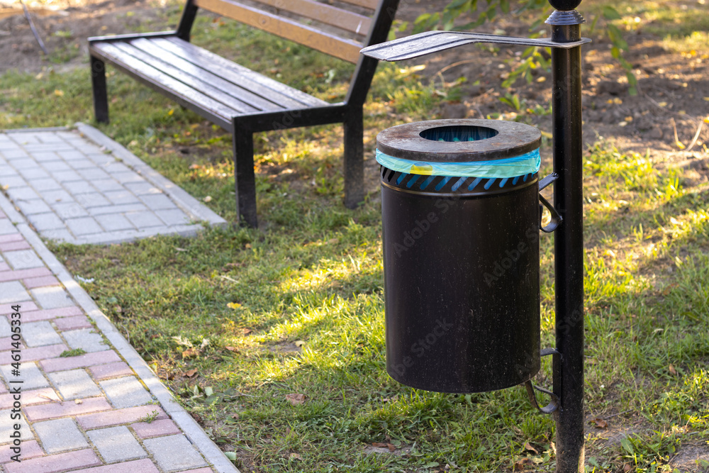 dirty trash can (bin) in the park with plastic bag inside,near a park brown bench.grass background. sidewalk.ecology concept.keep clean the environment. dirty trash can (bin) in the park with plastic bag inside,near a park brown bench.grass background. sidewalk.ecology concept.keep clean the environment.