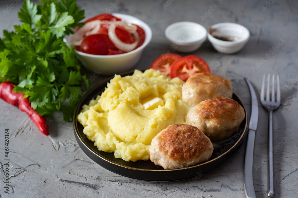 A hearty lunch: mashed potatoes, pork cutlets, tomato slices in a plate, tomato salad in a white bowl, fresh greens on a gray background. Close-up A hearty lunch: mashed potatoes, pork cutlets, tomato slices in a plate, tomato salad in a white bowl, fresh greens on a gray background. Close-up