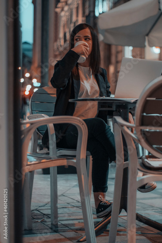 Directiva morena sentada en una cafeteria tomando café por la noche mientras completa sus tareas y trabajos de oficina en la ciudad con su laptop
