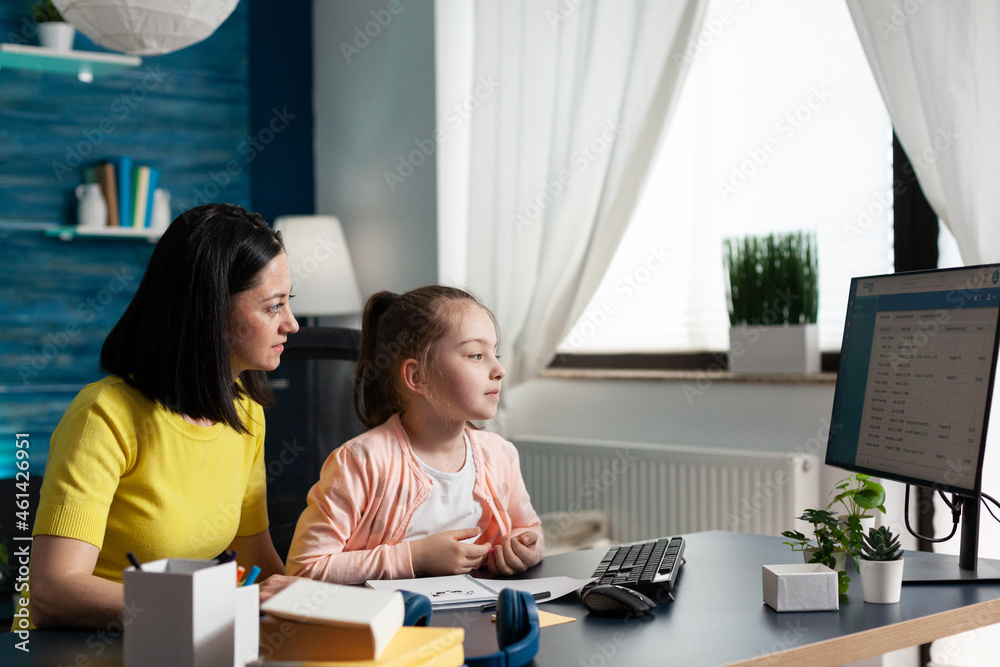 Mother sitting beside daughter doing school homework together analyzing elementary mathematics course on computer. Clever child studying literature during home schooling in living rom Mother sitting beside daughter doing school homework together analyzing elementary mathematics course on computer. Clever child studying literature during home schooling in living rom