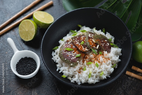 Black bowl with rice, smoked tuna fillet, green onion and sesame seeds, horizontal shot on a dark brown stone background