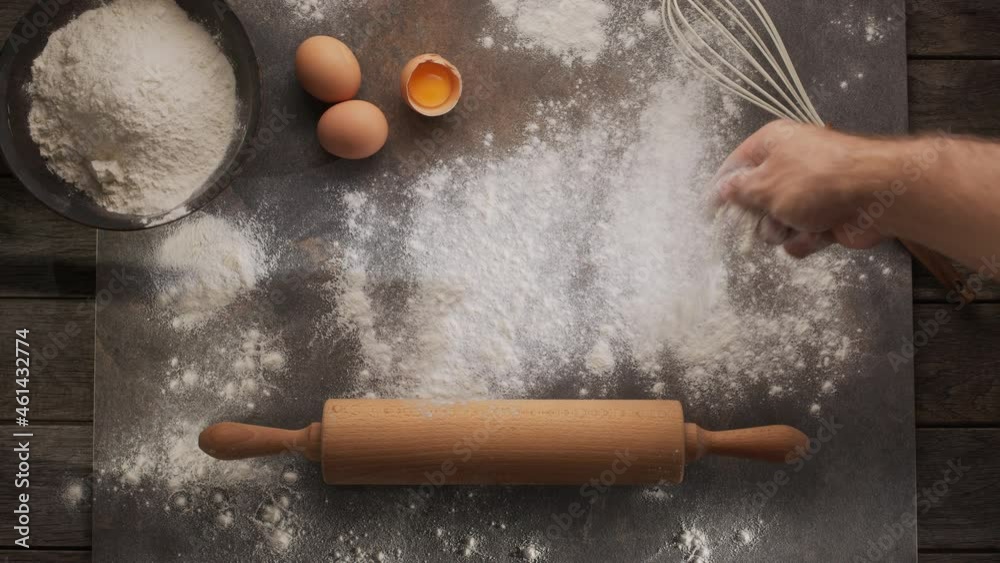 Chef's hand scatters flour on the table for rolling out the dough against the background of a rolling pin, eggs and a whisk. Home bakery concept. 