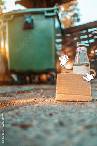 The concept of environmental pollution and waste sorting. Glass bottle with cartoon hands holding a empty cardboard box. Green garbage can in the background. Vertical orientation