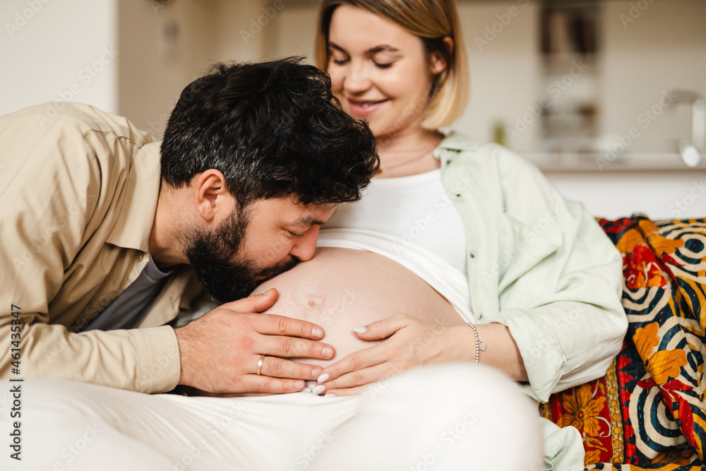 White man kissing his pregnant wife's belly while sitting on couch White man kissing his pregnant wife's belly while sitting on couch
