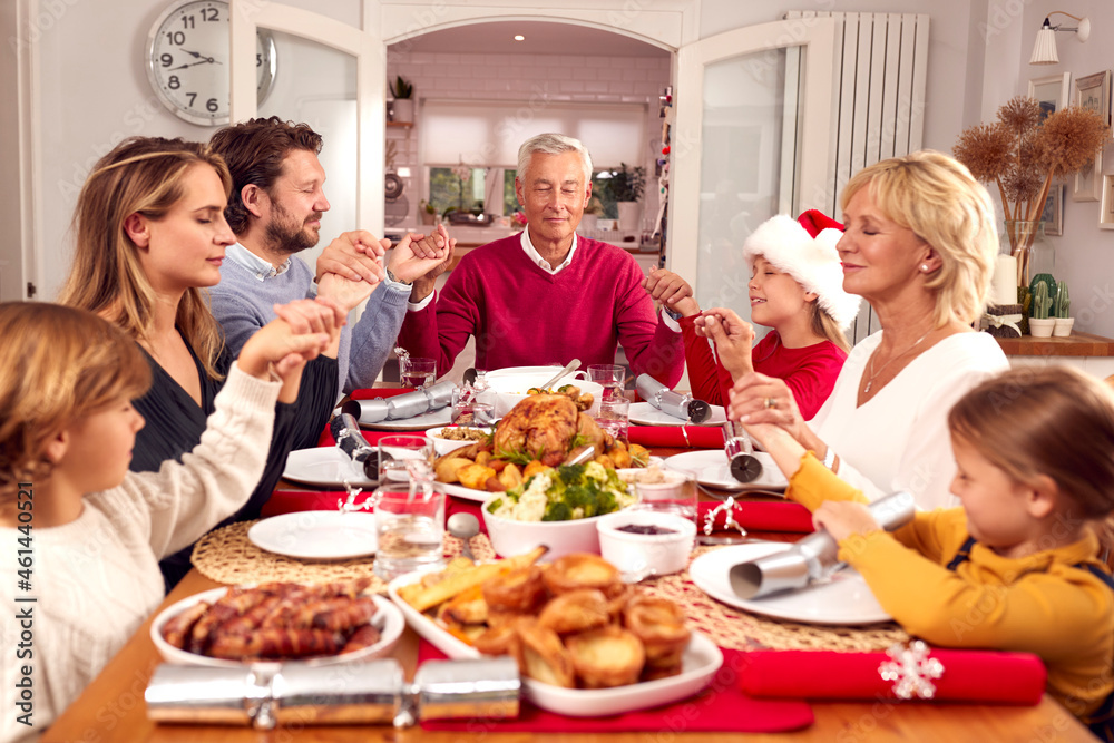 Multi Generation Family Hold Hands Around Table At Home Saying Grace Before Eating Christmas Meal