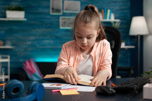 Schoolchild sitting at desk in living room holding school book reading educational literature story for online course during coronavirus quarantine. Reader child studying for academic exam