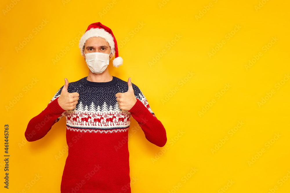 man in Christmas sweater with reindeer, medical mask, New Year's hat, a Santa Claus hat, gives a thumbs up, likes, OK sign. isolated yellow background with space for text. holiday concept, gifts, sale