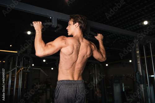 back view of shirtless muscular man working out on horizontal bar in gym.