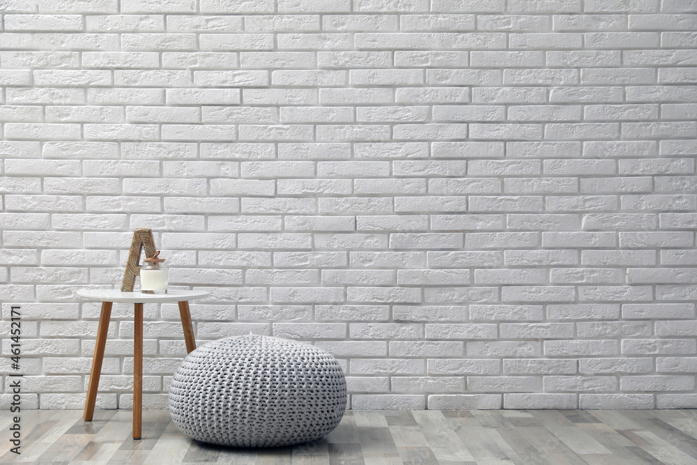 Comfortable knitted pouf, table and decor elements near white brick wall indoors, space for text. Interior design - obrazy, fototapety, plakaty 