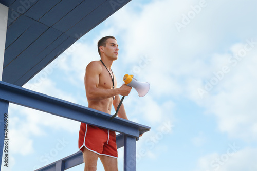 Handsome lifeguard with megaphone on watch tower against sky
