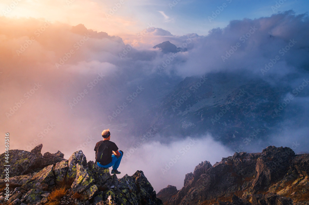 Man sits alone on the peak of rock. Hiker watching to autumn Sun at horizon . Beautiful moment the miracle of nature. Colorful mist in valley. Man hike. Person silhouette stand